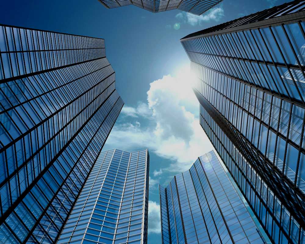 Upward view of glass skyscrapers against blue sky with clouds and sun flare, cool blue tones, sleek corporate mood