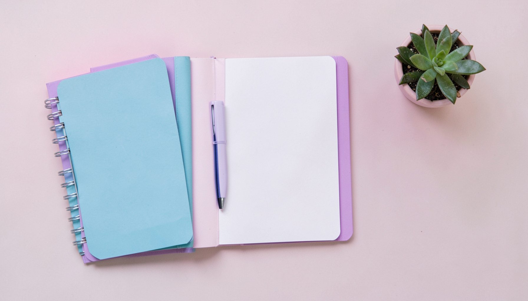 Overhead flat-lay of a tidy desk scene in soft pink, teal, and purple tones with a notebook, a pen, and a small potted plant